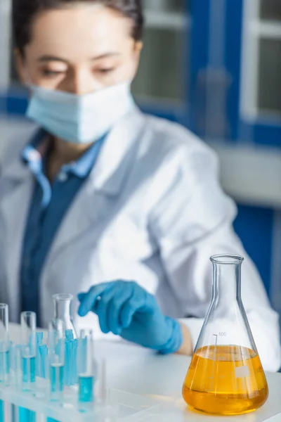 Selective focus of flask with yellow liquid near blurred bioengineer in medical mask — Stock Photo