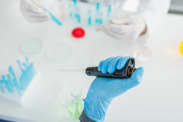 Abgeschnittene Ansicht eines Wissenschaftlers im Latexhandschuh, der eine Mikropipette in der Nähe eines verschwommenen Kollegen hält — Stockfoto