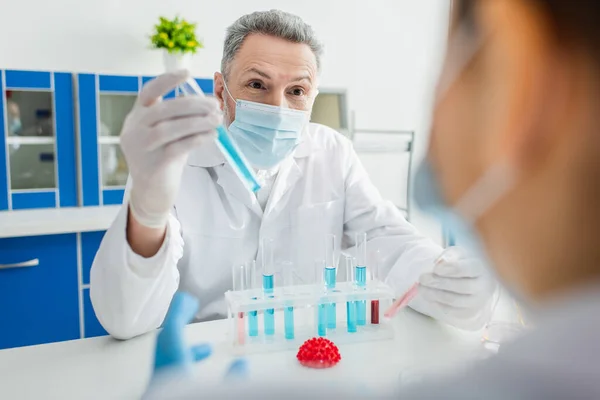 Bioingegnere in maschera medica che tiene la provetta vicino a collega offuscato e modello di batteri coronavirus — Foto stock