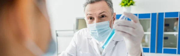 Cientista em máscara médica segurando tubo de ensaio perto colega borrado, banner — Fotografia de Stock