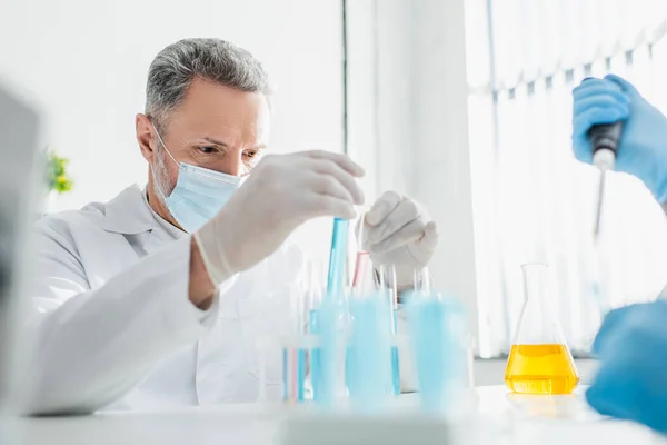 Bioengenheiro em máscara médica segurando tubos de ensaio em laboratório — Fotografia de Stock
