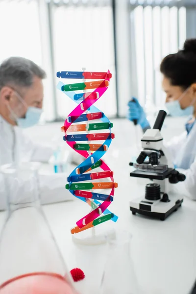 Selective focus of dna structure near microscope and geneticists in medical masks on blurred background — Stock Photo