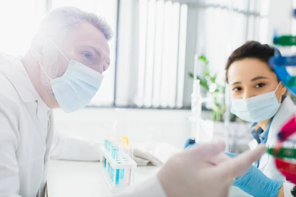 Mature geneticist in medical mask pointing at blurred dna model while working with colleague in lab — Stock Photo