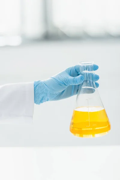 Visão parcial do cientista em luva de látex azul segurando frasco com líquido amarelo — Fotografia de Stock