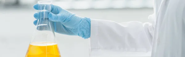 Partial view of scientist in latex glove holding medical flask with yellow liquid, banner — Stock Photo