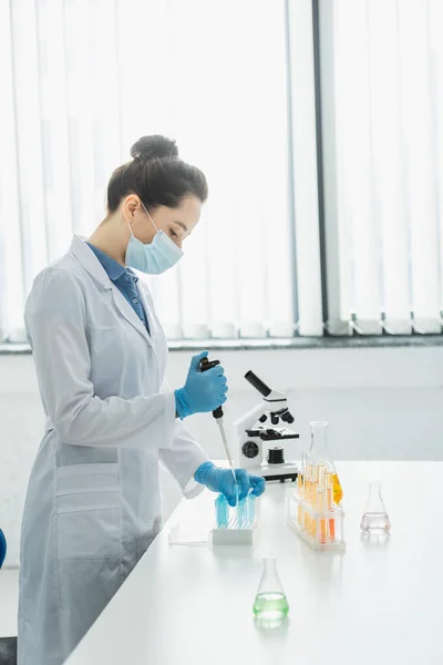 Visão lateral do cientista na máscara médica que trabalha com micropipeta e tubos de ensaio perto do microscópio — Fotografia de Stock
