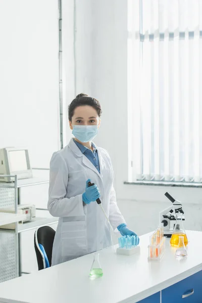 Biologe in medizinischer Maske blickt in Kamera, während er Mikropipette in der Nähe von Reagenzgläsern hält — Stockfoto
