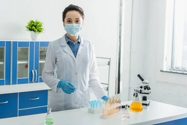 Bioingeniero en máscara médica mirando la cámara cerca de tubos de ensayo, microscopio y frascos - foto de stock