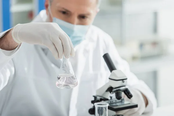 Blurred biologist in medical mask holding flask with liquid near microscope — Stock Photo