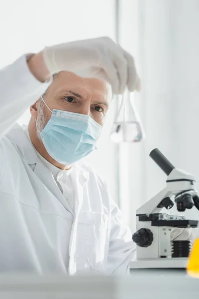 Middle aged scientist in medical mask holding blurred flask near microscope — Stock Photo