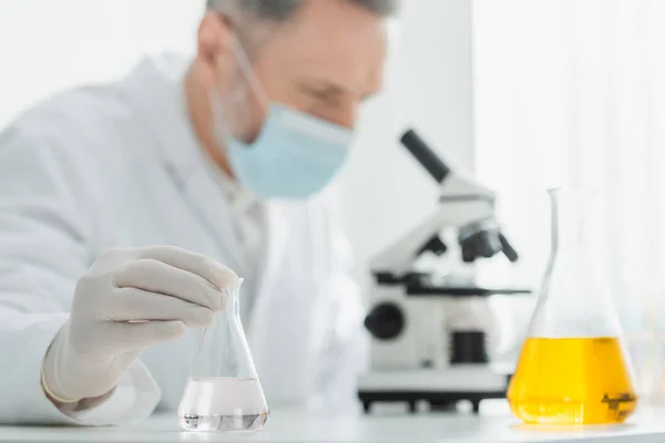 Blurred biotechnologist in medical mask looking in microscope near flasks with liquid — Stock Photo