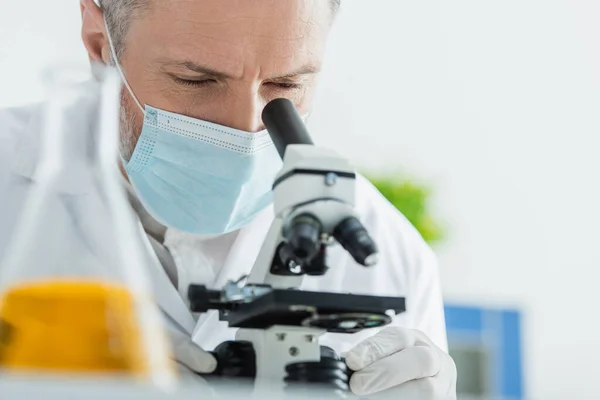 Bioingeniero maduro en máscara médica que trabaja con microscopio en laboratorio químico - foto de stock