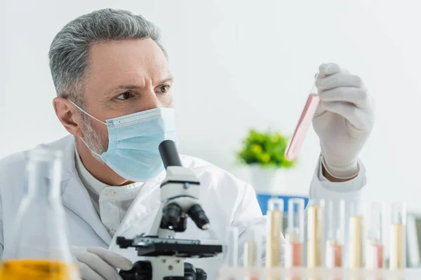 Grey-haired bioengineer in medical mask looking at test tube near microscope — Stock Photo