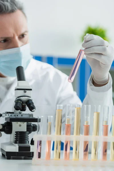Размытый биоинженер в медицинской маске держит пробирку с жидкостью возле микроскопа — стоковое фото