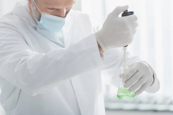 Scientist in medical mask and latex gloves holding micropipette and flask with liquid — Stock Photo