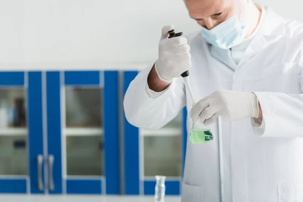 Scientifique en masque médical travaillant avec micropipette et fiole avec liquide en laboratoire — Photo de stock