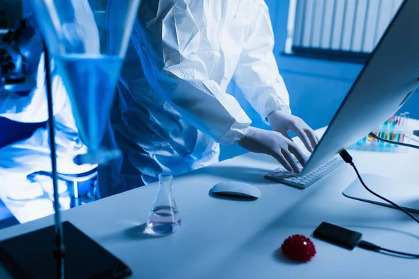 Cropped view of virologist in latex gloves typing on keyboard near flask with liquid — Stock Photo