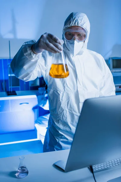 Cientista de fato hazmat segurando frasco com líquido laranja perto monitor de computador borrado em laboratório — Fotografia de Stock