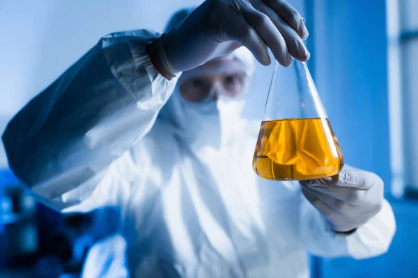 Blurred biotechnologist in hazmat suit holding flask with orange liquid — Stock Photo