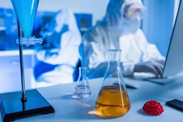 Flasks with liquid and coronavirus bacteria model near blurred bioengineers in hazmat suits — Stock Photo