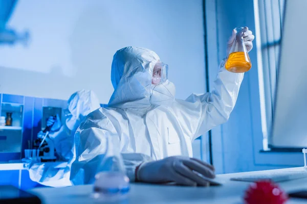 Биоинженер в защитном костюме смотрит на фляжку с оранжевой жидкостью во фляжке — стоковое фото