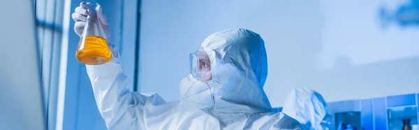 Virólogo en traje de materiales peligrosos, gafas y máscara médica que sostiene el frasco con líquido naranja en el laboratorio, pancarta - foto de stock