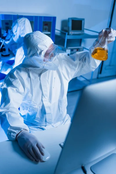 Científico en traje de materiales peligrosos que sostiene el frasco con líquido naranja cerca del monitor borroso en el laboratorio - foto de stock