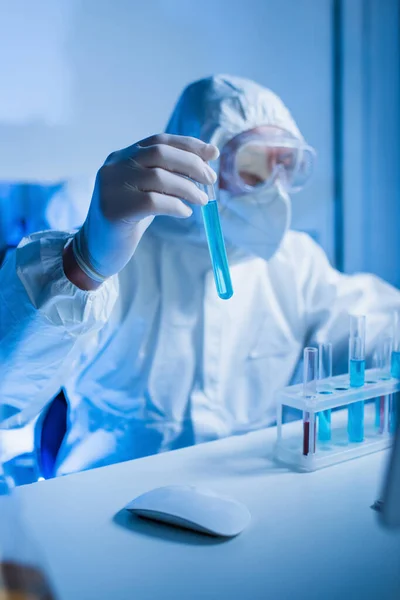 Blurred virologist in hazmat suit and goggles holding test tube with blue liquid — Stock Photo