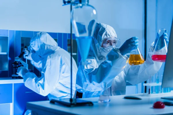 Генетик в индивидуальном защитном оборудовании держит колбы с красочной жидкостью в лаборатории — стоковое фото