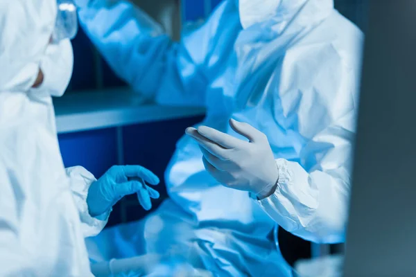 Vue recadrée de scientifiques flous en combinaison de matières dangereuses et gants en latex travaillant en laboratoire — Photo de stock