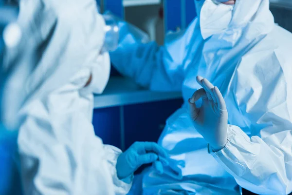 Vista recortada del virólogo en traje de hazmat y guantes de látex mostrando gesto bien cerca de colega - foto de stock