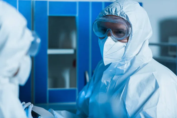 Bio-ingénieur en combinaison Hazmat, lunettes et masque médical en regardant son collègue au premier plan flou — Photo de stock