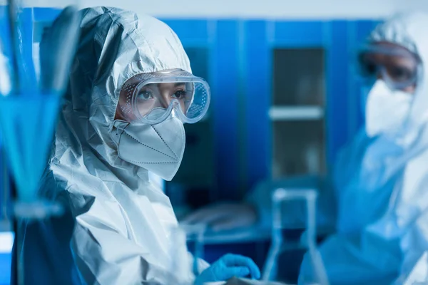 Mulher em óculos, máscara médica e terno hazmat perto cientista em fundo borrado — Fotografia de Stock