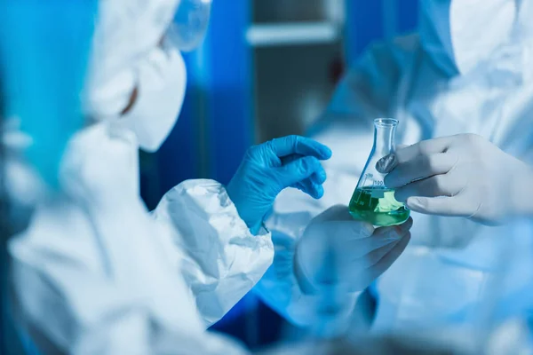 Обрезанный вид биоинженера в латексных перчатках, держащего фляжку с жидкостью рядом с размытым ученым — стоковое фото