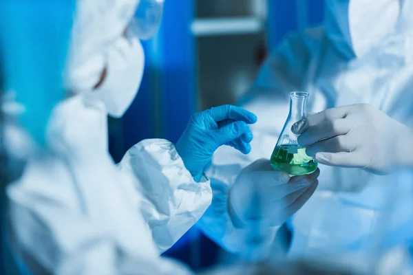 Vista recortada de bioingeniero en guantes de látex sosteniendo matraz con líquido cerca de científico borroso - foto de stock