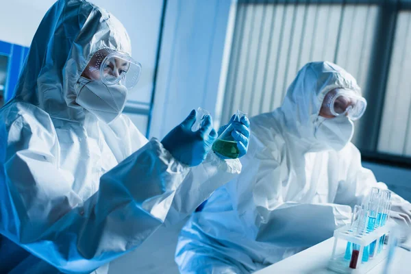 Científico en traje de materiales peligrosos sosteniendo frascos médicos mientras trabaja con un colega en el laboratorio - foto de stock