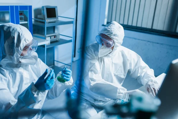 Bioingeniero con frascos mirando a su colega en traje de materiales peligrosos cerca de la computadora, borrosa primer plano - foto de stock