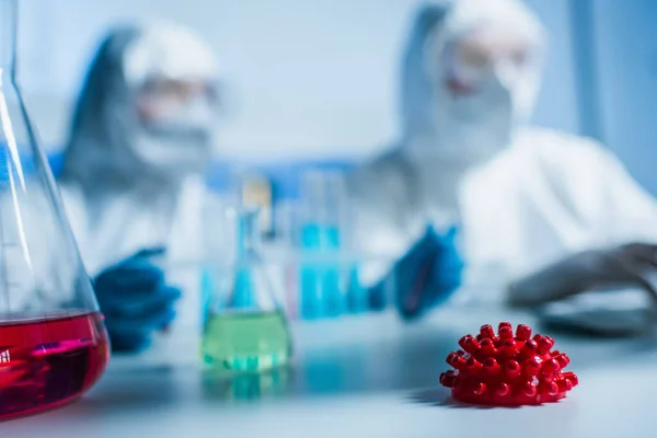 Enfoque selectivo del frasco con líquido rojo y el modelo de bacterias coronavirus cerca de virólogos borrosos - foto de stock