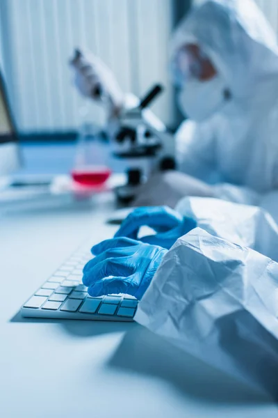 Scientist in latex gloves typing on computer keyboard while colleague working on blurred background — Stock Photo