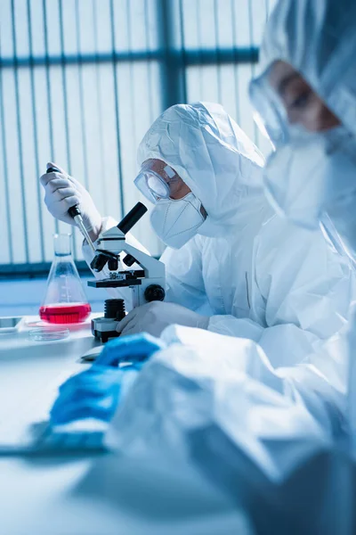 Vista lateral del bioingeniero en traje de materiales peligrosos que trabaja con microscopio y micropipeta cerca de colega borroso - foto de stock