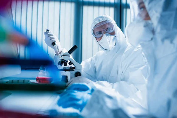 Hombre con gafas y máscara médica cerca del microscopio y científico en primer plano borrosa - foto de stock