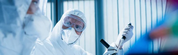 Científico en gafas y máscara médica cerca del microscopio y colega en primer plano borrosa, bandera - foto de stock