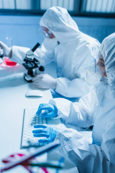 Frau in Schutzanzug tippt auf Computertastatur nahe verschwommenem Wissenschaftler mit Mikroskop — Stockfoto