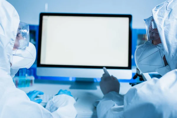 Scientist in hazmat suit and medical mask pointing at monitor with white screen while working with colleague — Stock Photo