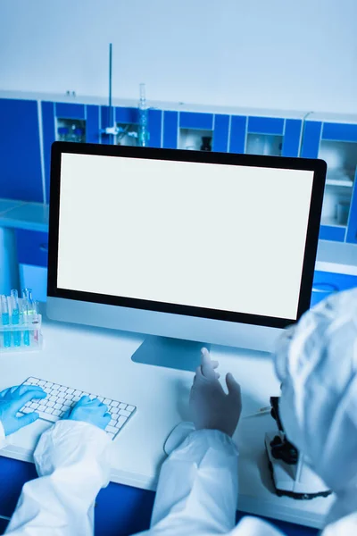 Vista ritagliata dello scienziato in guanti di lattice che punta al monitor con schermo bianco mentre il collega digita sulla tastiera — Foto stock
