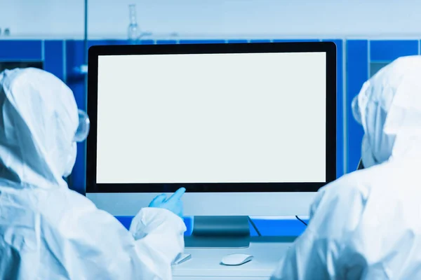 Visão traseira do cientista em terno hazmat apontando para monitor com tela branca perto colega — Fotografia de Stock