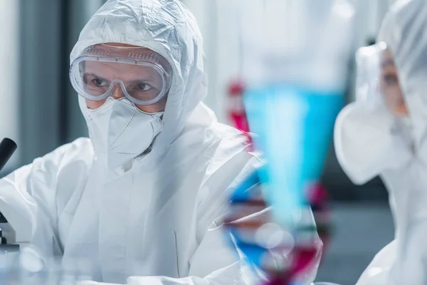 Mann mit Schutzbrille und Schutzanzug arbeitet neben verschwommenem Kollegen im Labor — Stockfoto