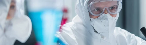 Científico en traje de materiales peligrosos, gafas y máscara médica cerca de colega borrosa en el laboratorio, pancarta - foto de stock