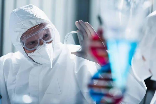 Virólogo en máscara médica y gafas que muestran placa de Petri a colega borrosa - foto de stock