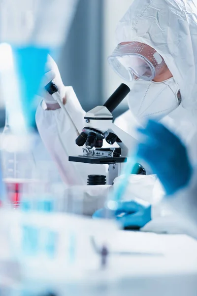 Bioengineer in hazmat suit working with microscope on blurred foreground — Stock Photo